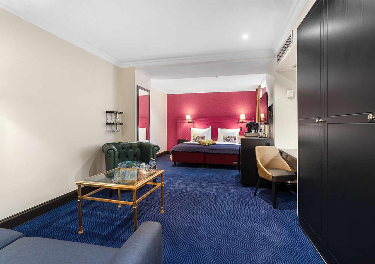 Una camera d'albergo con tappeto blu, una poltrona verde, un tavolino da caffè in vetro, un letto con biancheria rossa e un'armadio in legno scuro.