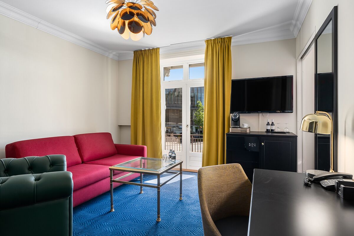 A modern hotel room with a red sofa, green armchair, and a glass coffee table. There is a black desk with a telephone and a lamp. A flat-screen TV is mounted on the wall, and a kitchen area is visible through the window.