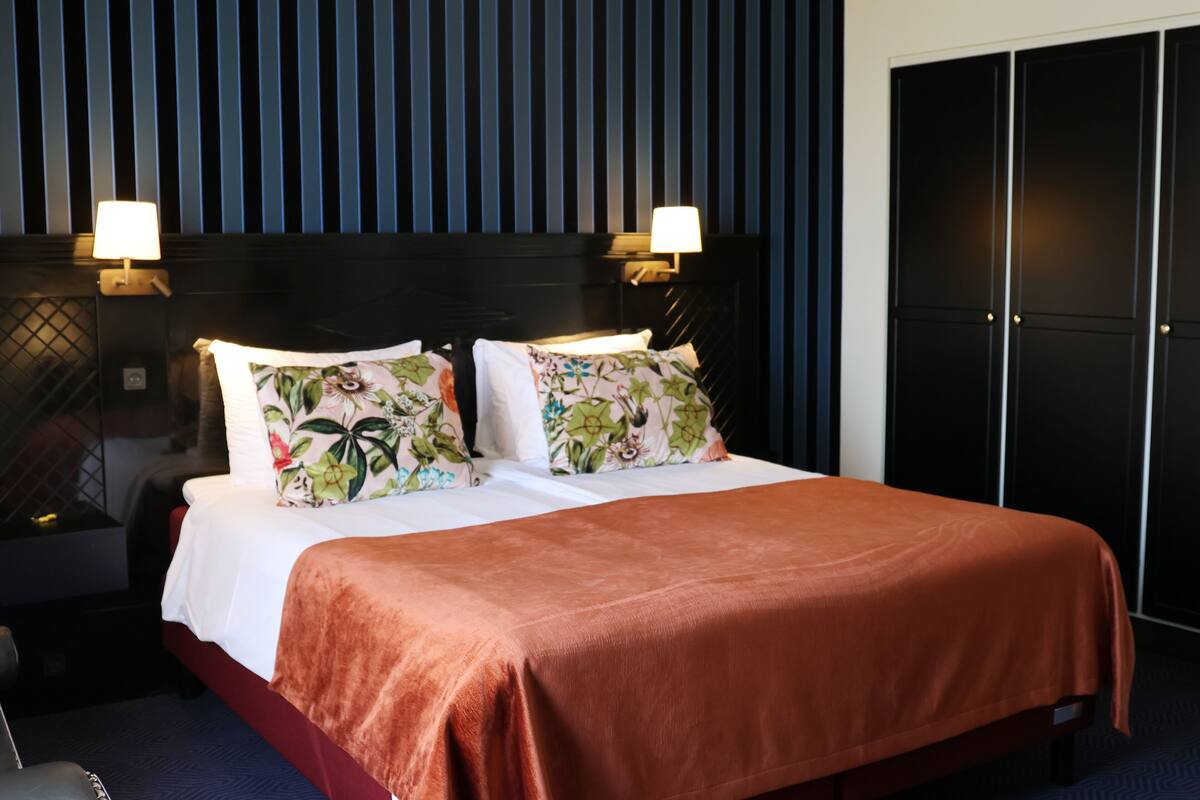 A hotel room with a large bed, two bedside lamps, a dark headboard, and a wardrobe.