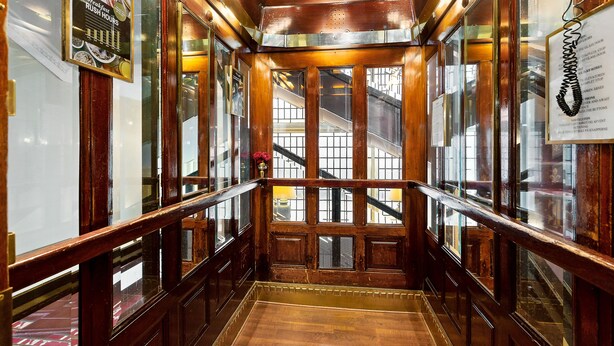 Interior de un ascensor de madera con puertas de vidrio y barandilla metálica.
