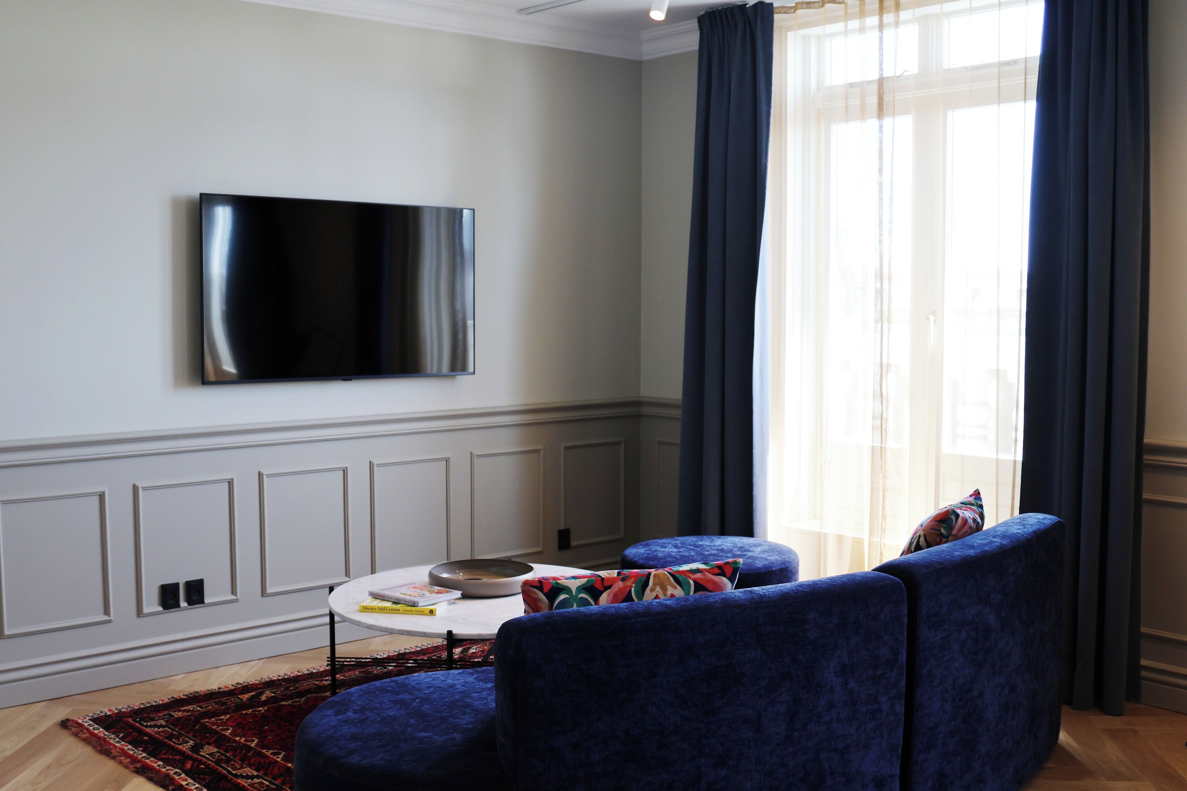 A modern living room with a flat-screen TV mounted on the wall, a round coffee table, two blue armchairs, and a patterned rug.
