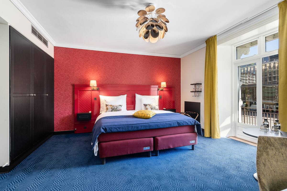A hotel room with a large bed, red walls, a blue carpet, and a view of the city through the window.