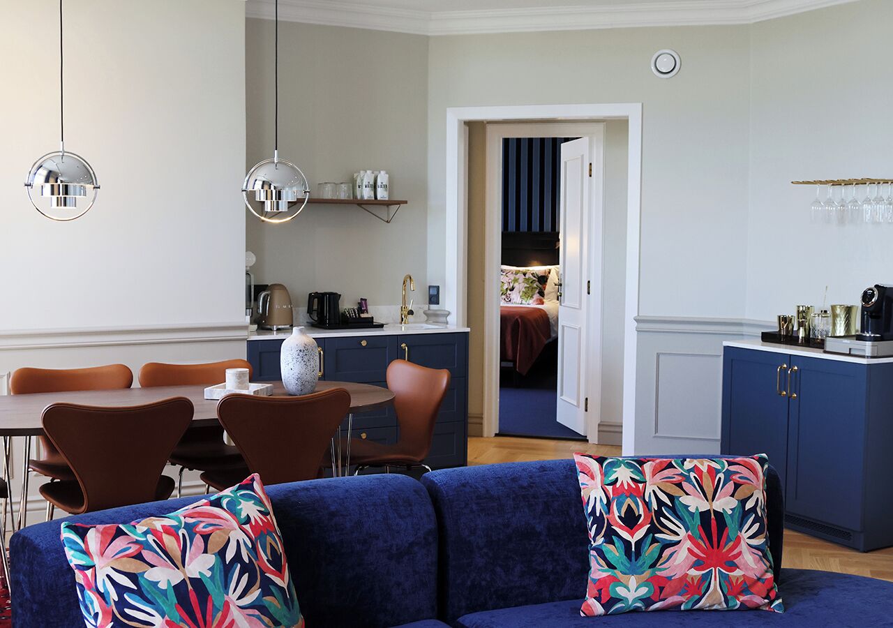 A modern living room with a blue sofa, a dining area with brown chairs, and a kitchen with blue cabinets.