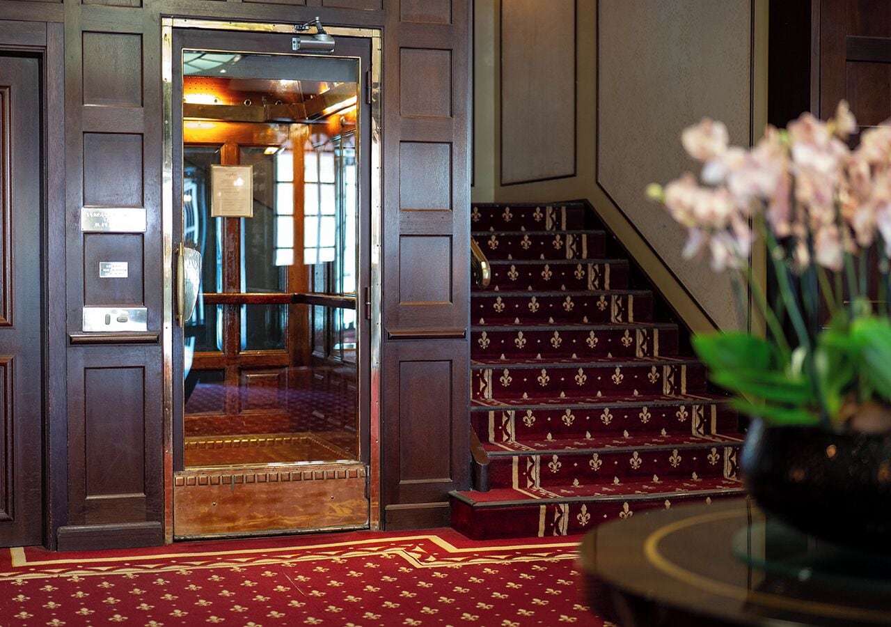 A hotel lobby with a grand wooden door, a red carpeted staircase, and a potted plant.
