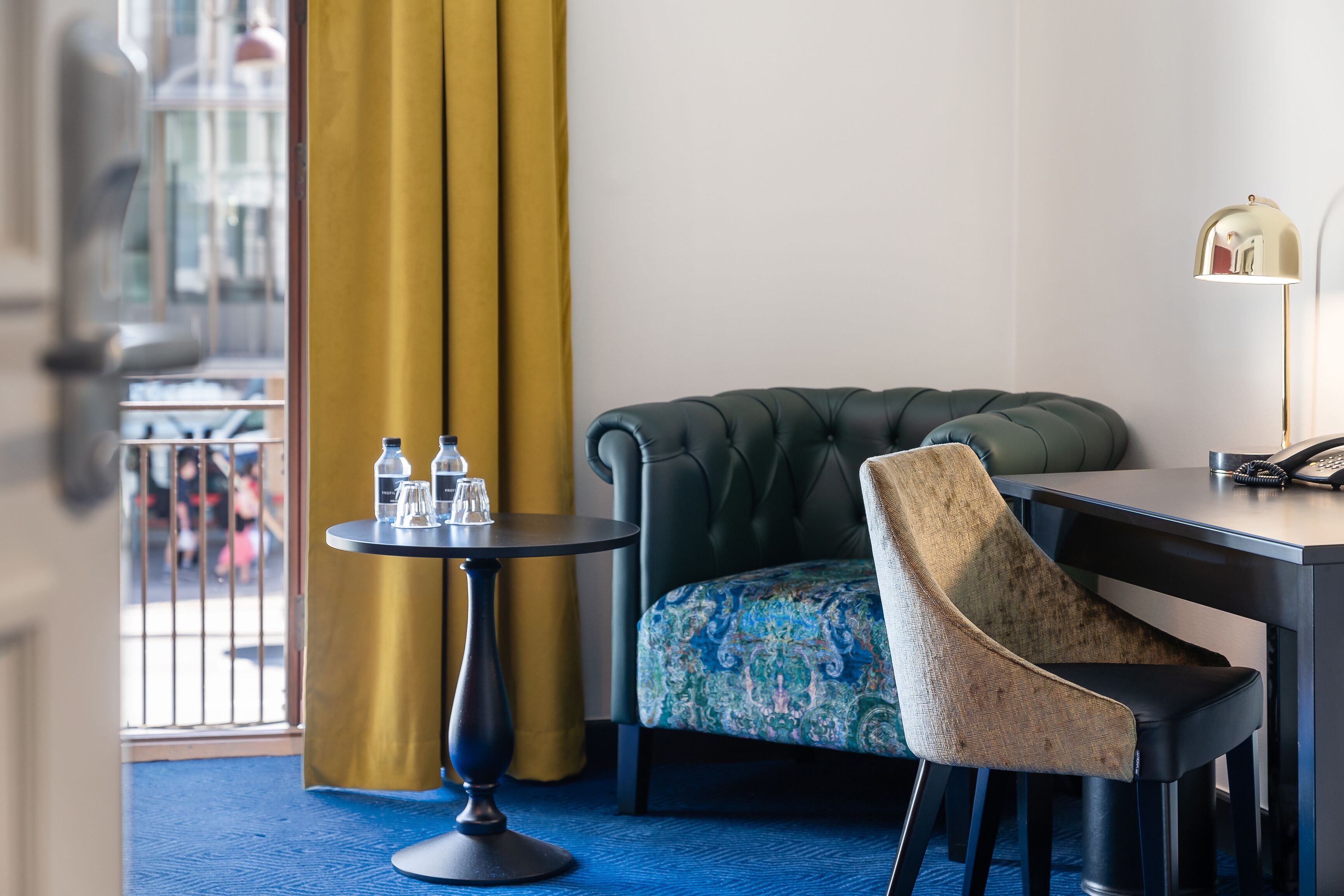 A hotel room with a tufted armchair, a small round table with a lamp, and a patterned sofa.
