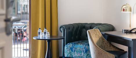 A hotel room with a tufted armchair, a small round table with a lamp, and a patterned sofa.