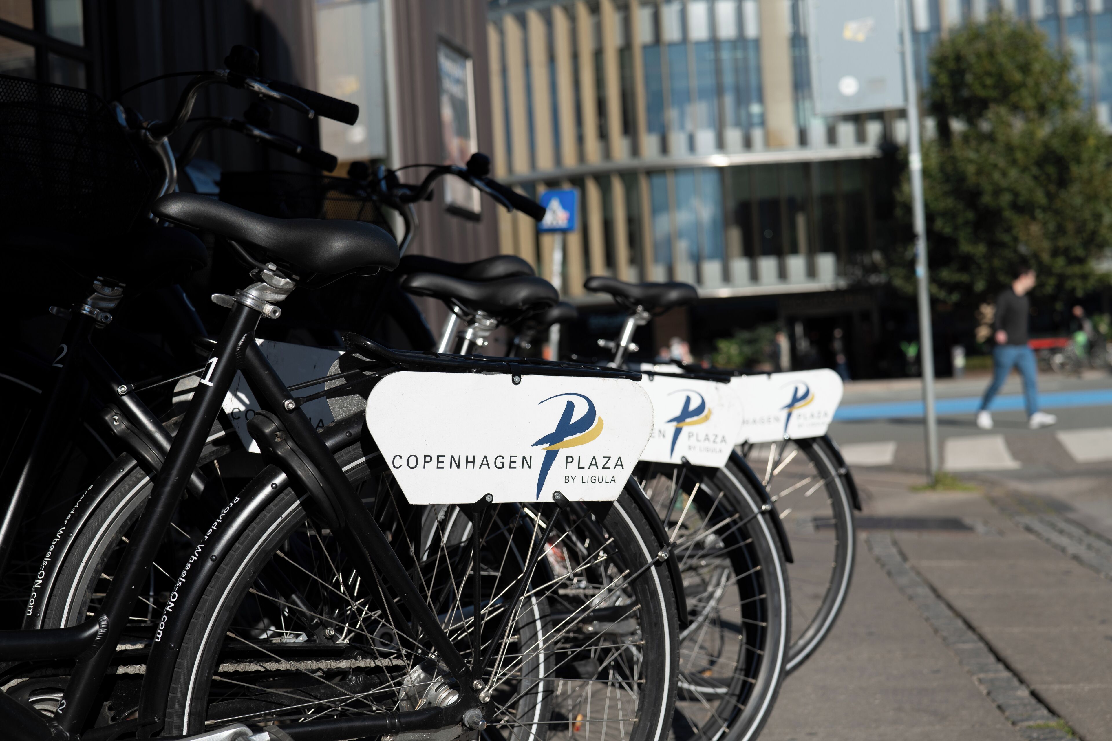 Una hilera de bicicletas con letreros que dicen 'Copenhagen Plaza by Ligula', estacionadas en una acera.