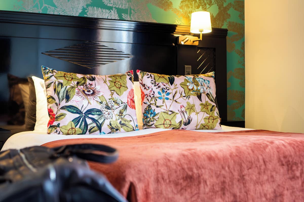 A bed with floral-patterned pillows, a wooden headboard, and a wall with a leafy pattern.