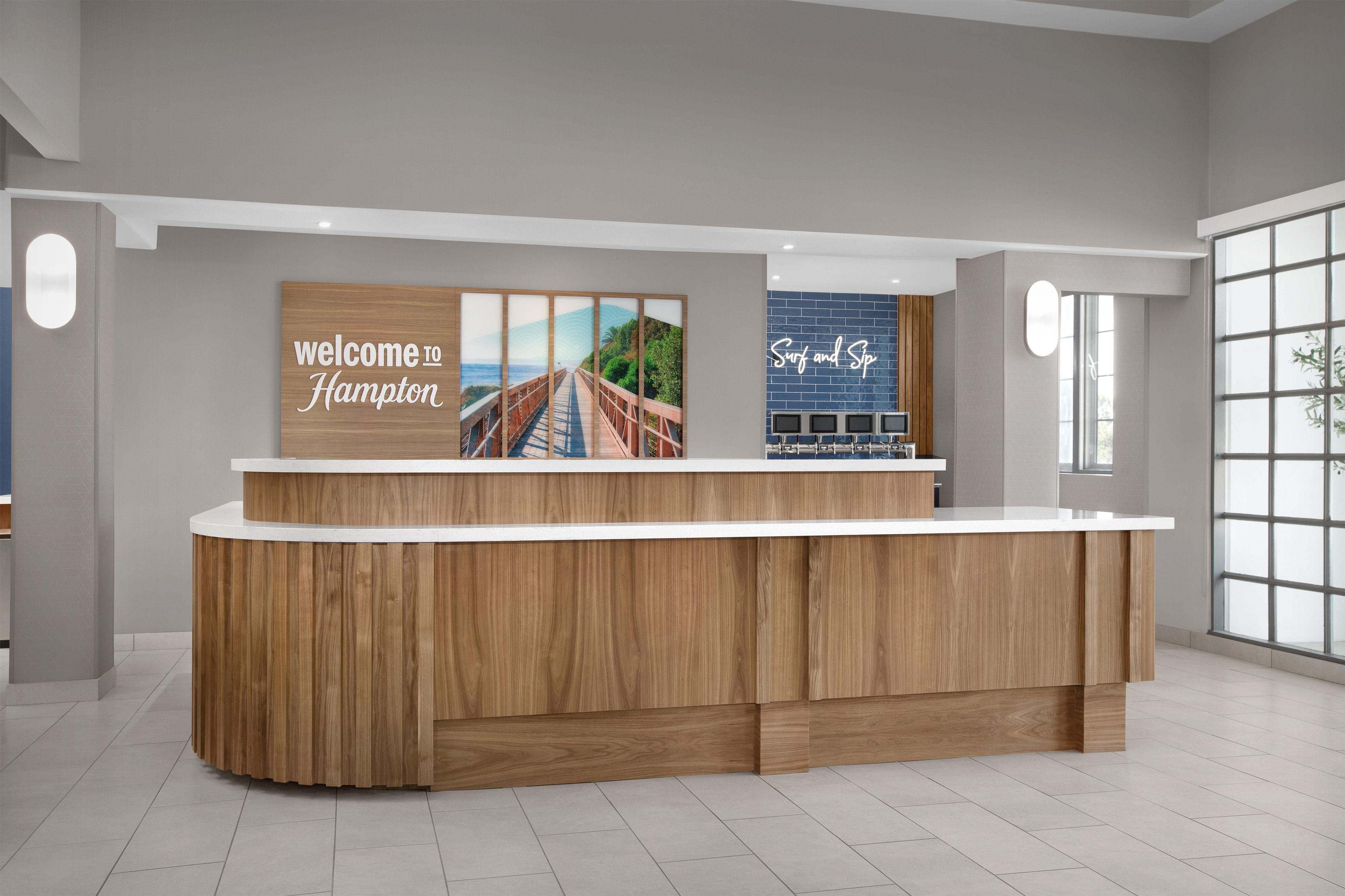 A reception desk with a wooden finish, a 'Welcome to Hampton' sign, and a 'Surf and Sip' sign.