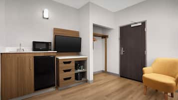 Una habitación compacta con kitchenette, televisor de pantalla plana, armario de madera, pequeño refrigerador, microondas, silla de madera y puerta oscura.