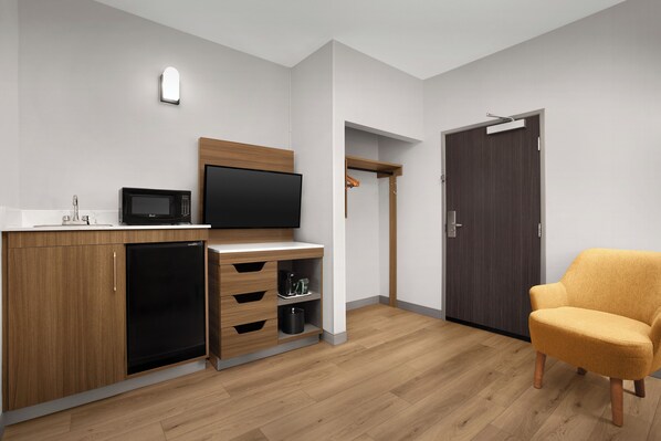 A compact room with a kitchenette, a flat-screen TV, a wooden cabinet, a small refrigerator, a microwave, a wooden chair, and a dark door.