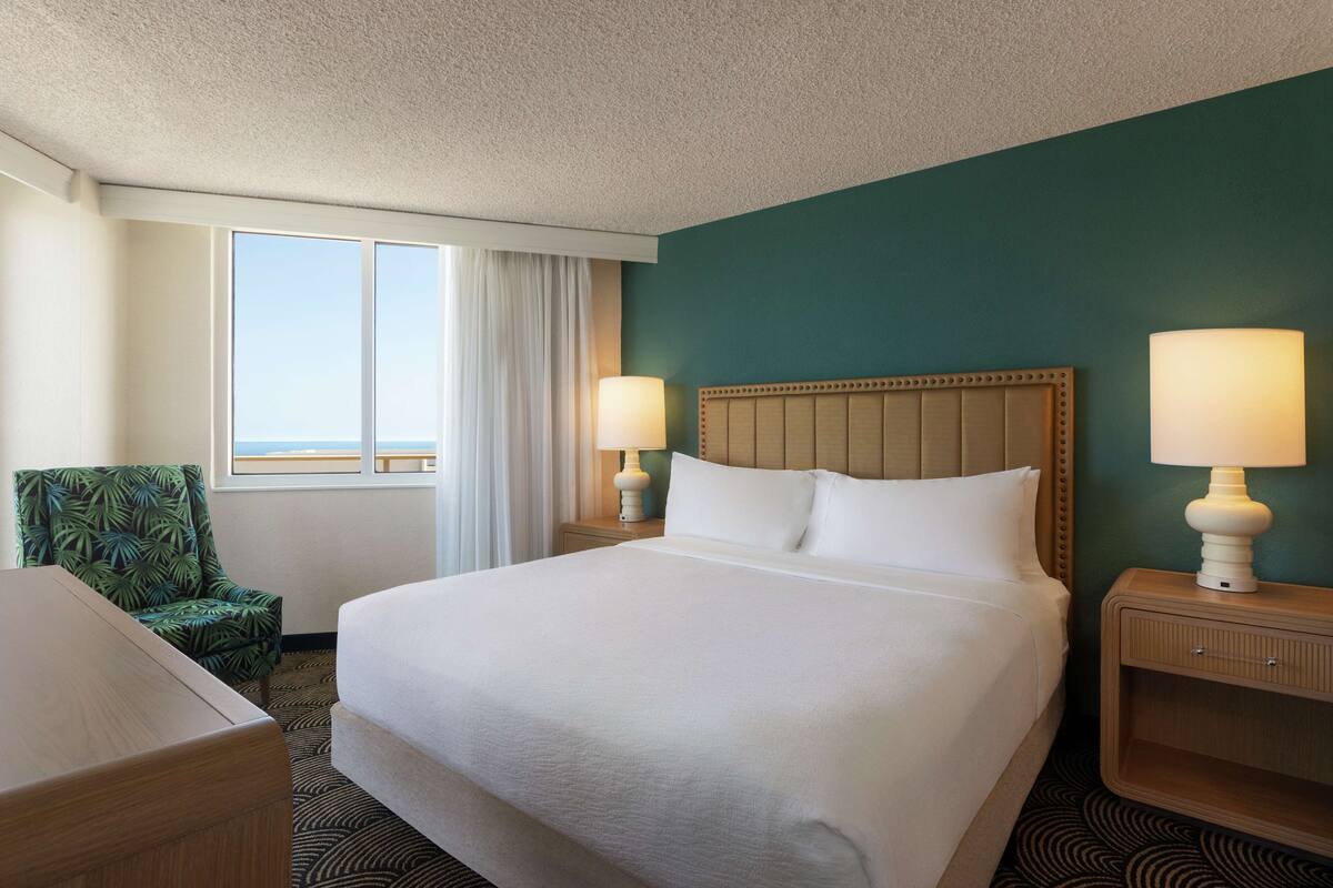 Ein Hotelzimmer mit einem großen Bett, zwei Nachttischlampen, einem Sessel, einem Nachttisch und Blick aufs Meer.