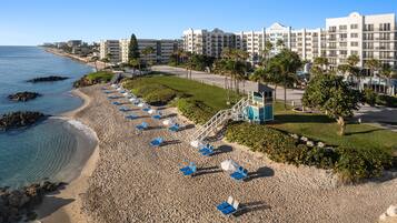 Una playa con hileras de sillas azules y sombrillas, una torre de salvavidas y edificios de hotel al fondo.