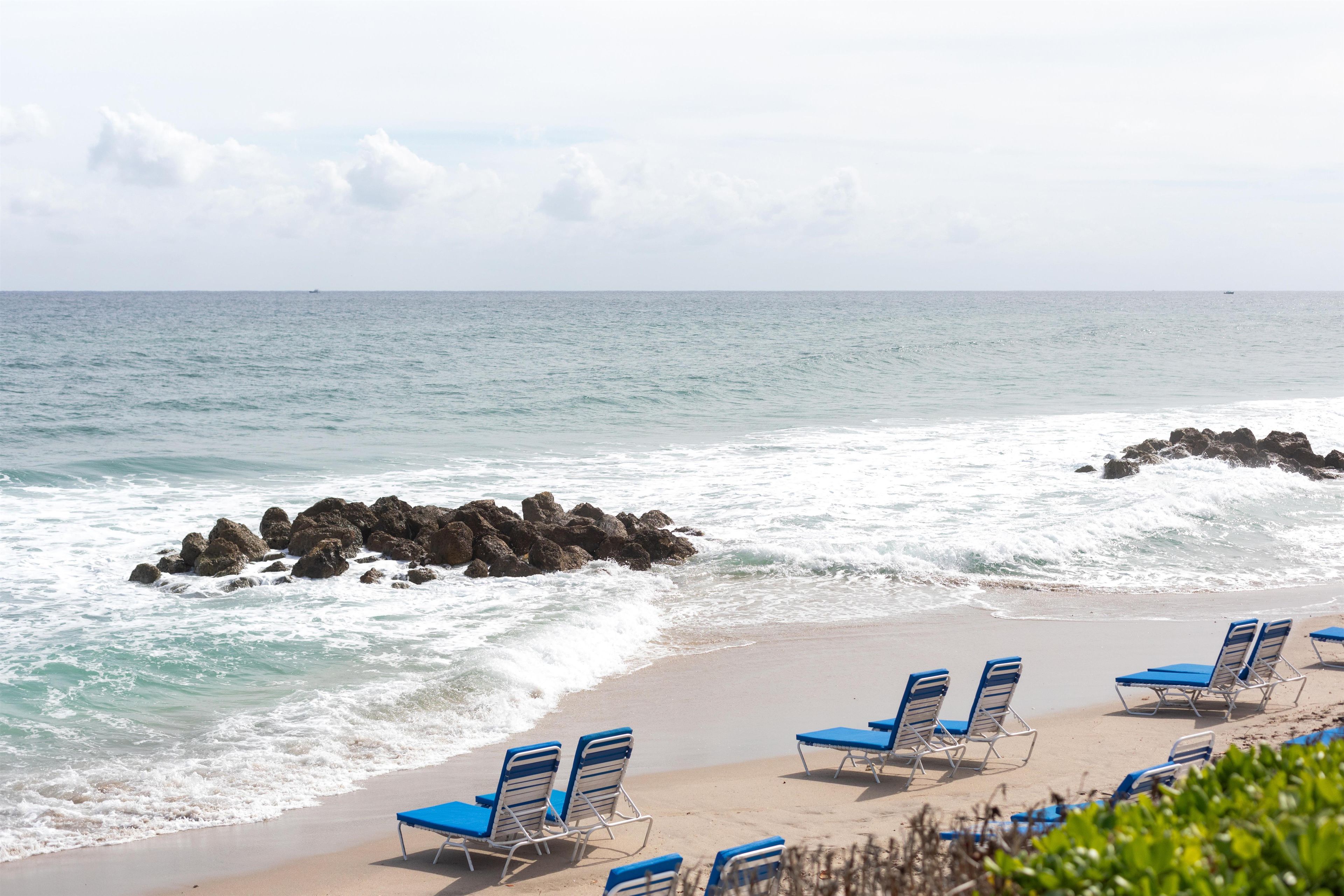 A beach with waves crashing onto rocks and a row of blue beach chairs.