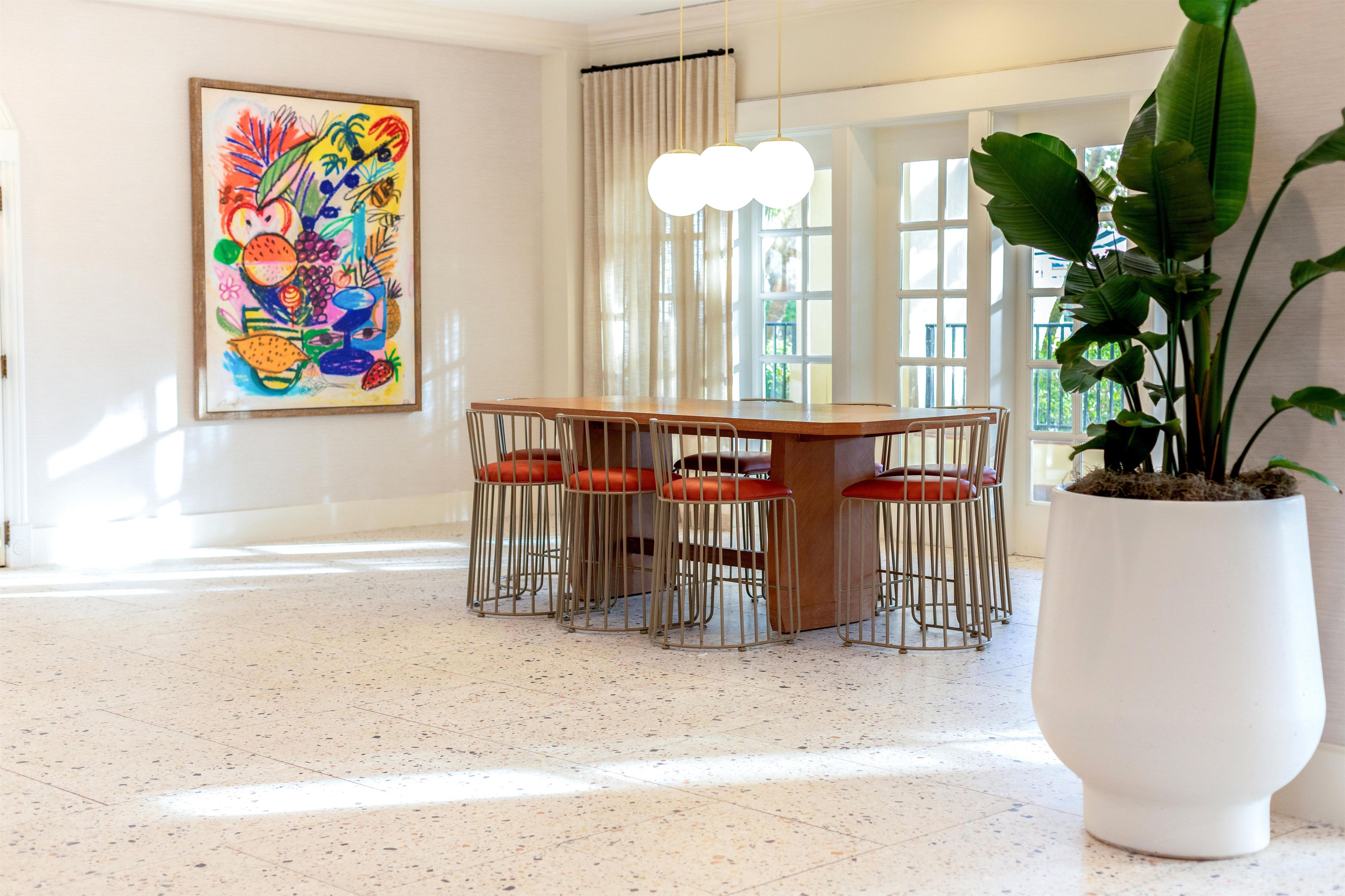 A dining area with a wooden table and metal chairs, a large potted plant, and a colorful abstract painting on the wall.