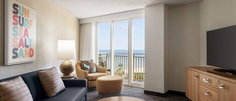 Habitación de hotel con sofá, reposapiés y televisor sobre un mueble de madera. Amplia ventana con vistas al océano.