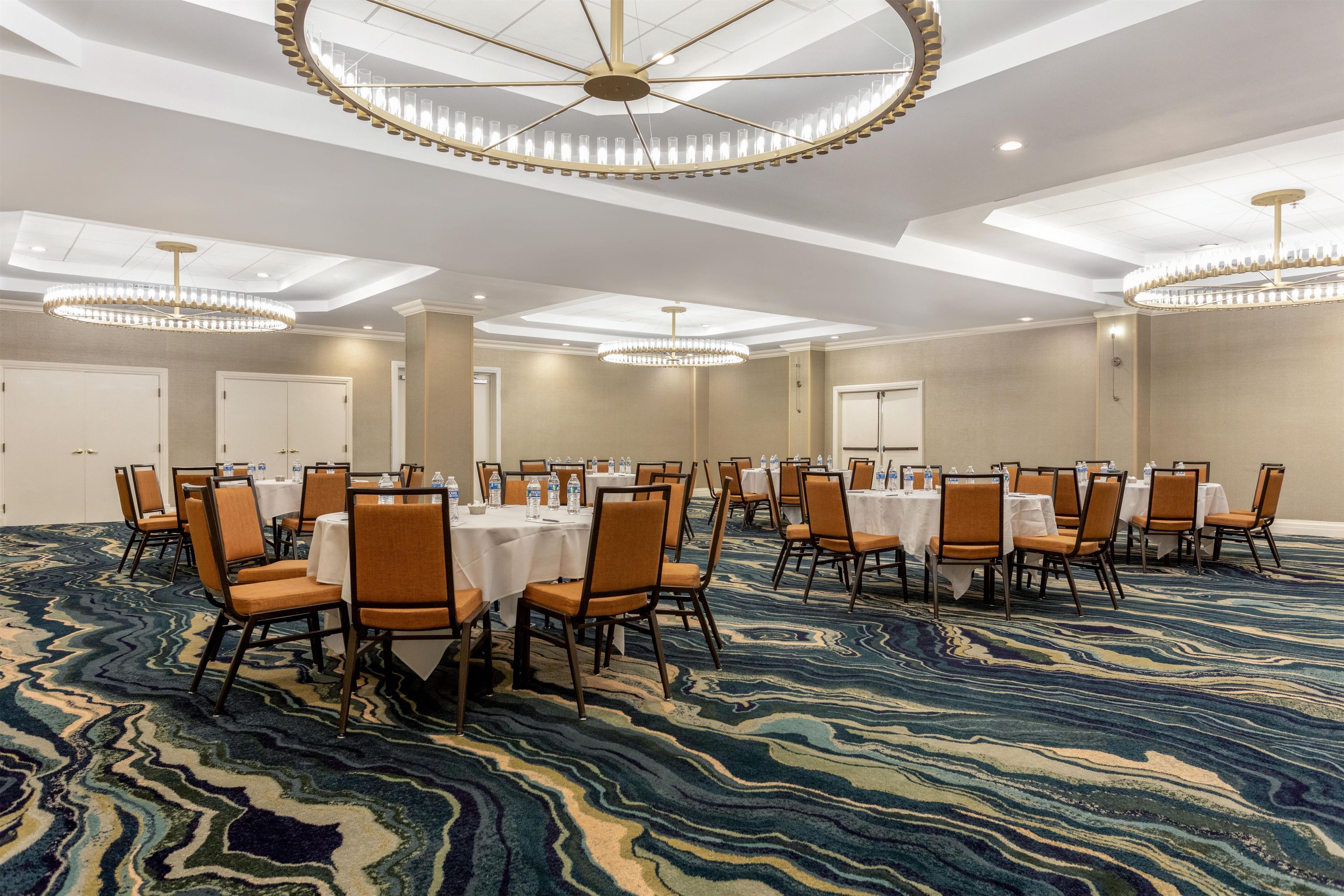 Un salón de conferencias con mesas y sillas redondas, alfombra estampada y un candelabro circular en el techo.