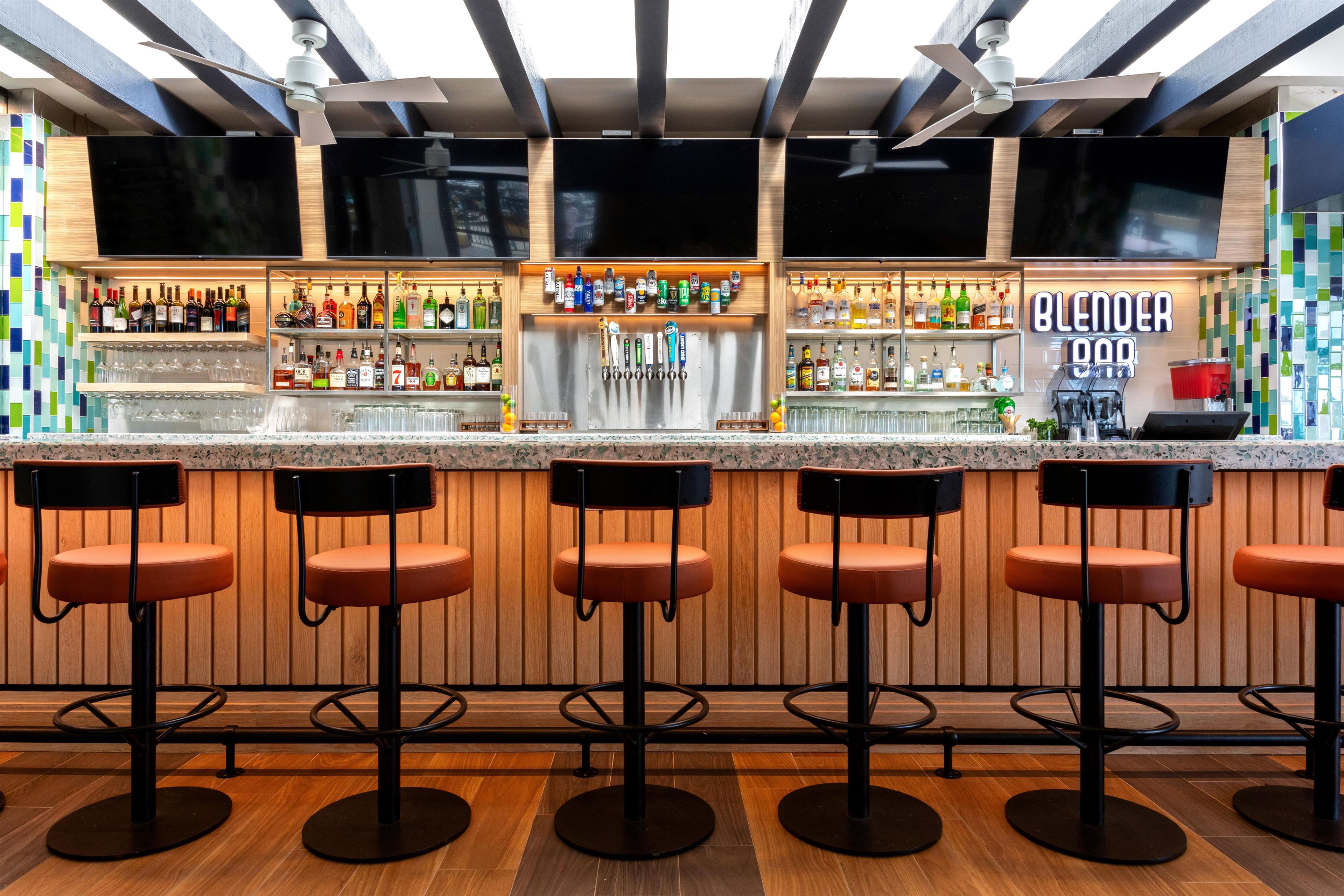 Un área de bar con varios taburetes, una barra con una variedad de botellas y un letrero que dice 'BLENDER BAR'.
