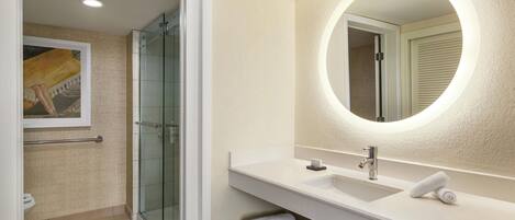 Un baño moderno con ducha de vidrio, lavamanos blanco y espejo redondo con luz.