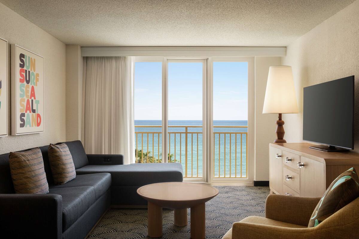 Ein Hotelzimmer mit einer Sitzecke, einem kleinen Tisch, einem Fernseher auf einer Holzkommode und einem großen Fenster mit Meerblick.