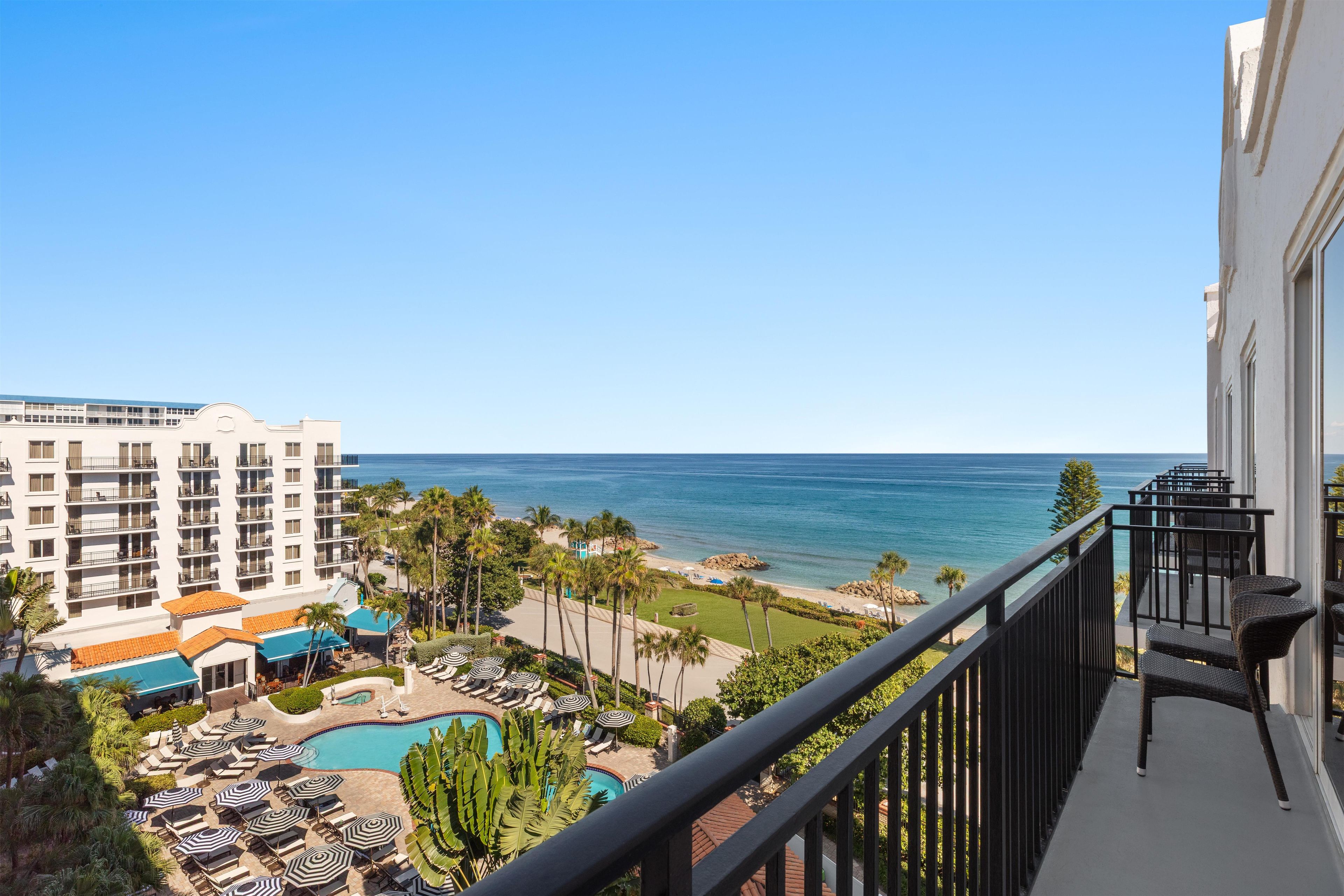A hotel balcony with a view of the ocean, a pool area with sun loungers, and a building with multiple floors.