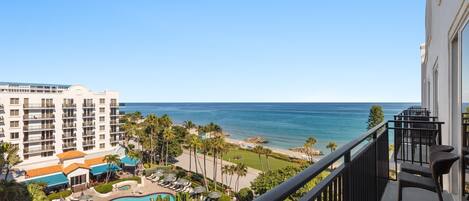 A hotel balcony with a view of the ocean, a pool area with sun loungers, and a building with multiple floors.