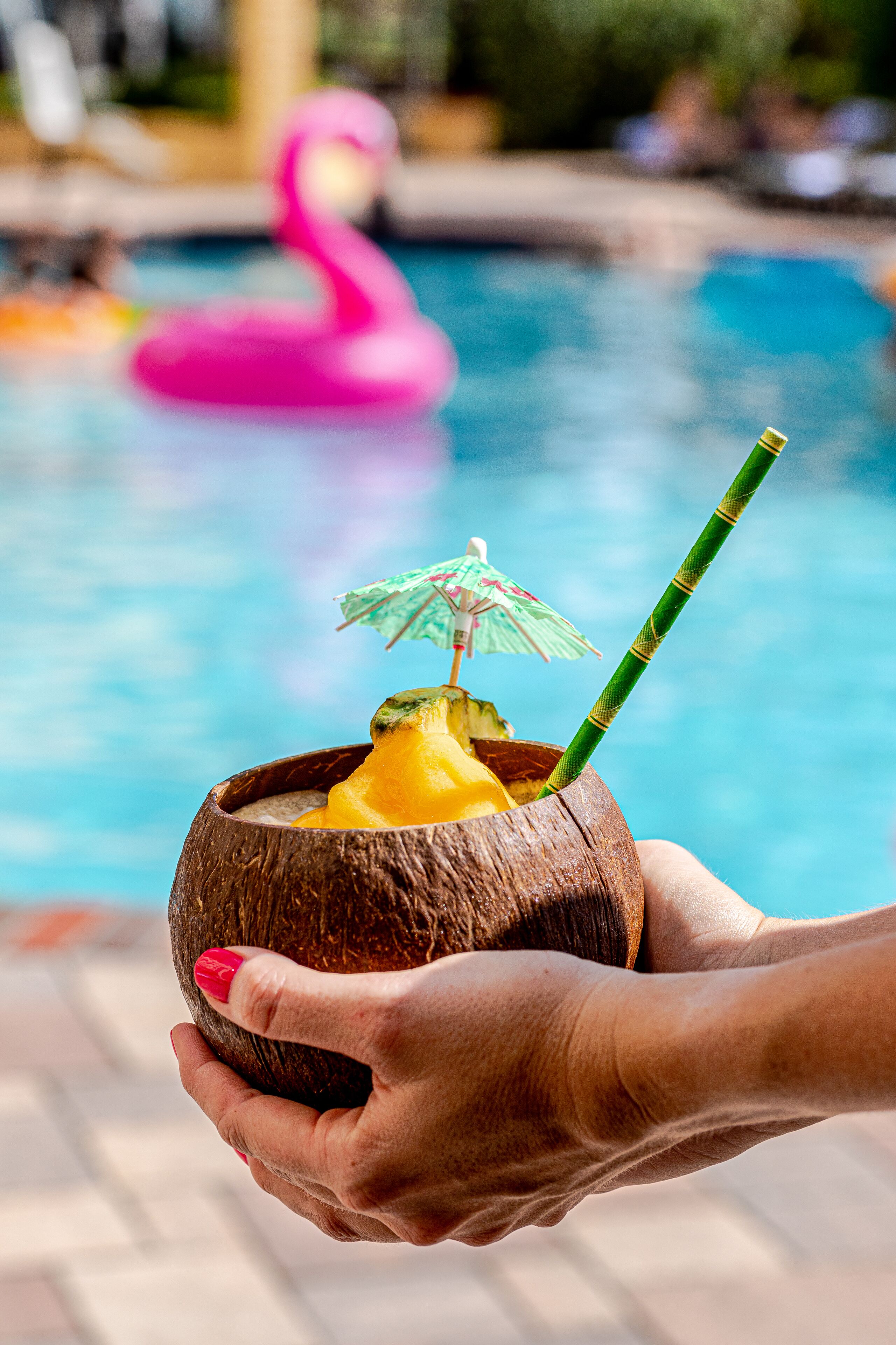 Una mano sostiene un coco con una pajita y un paraguas pequeño, con una piscina y un flotador con forma de flamenco rosa al fondo.