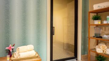 A bathroom with a glass shower door, a shelf with towels, a vase with a flower, and a shelf with toiletries.