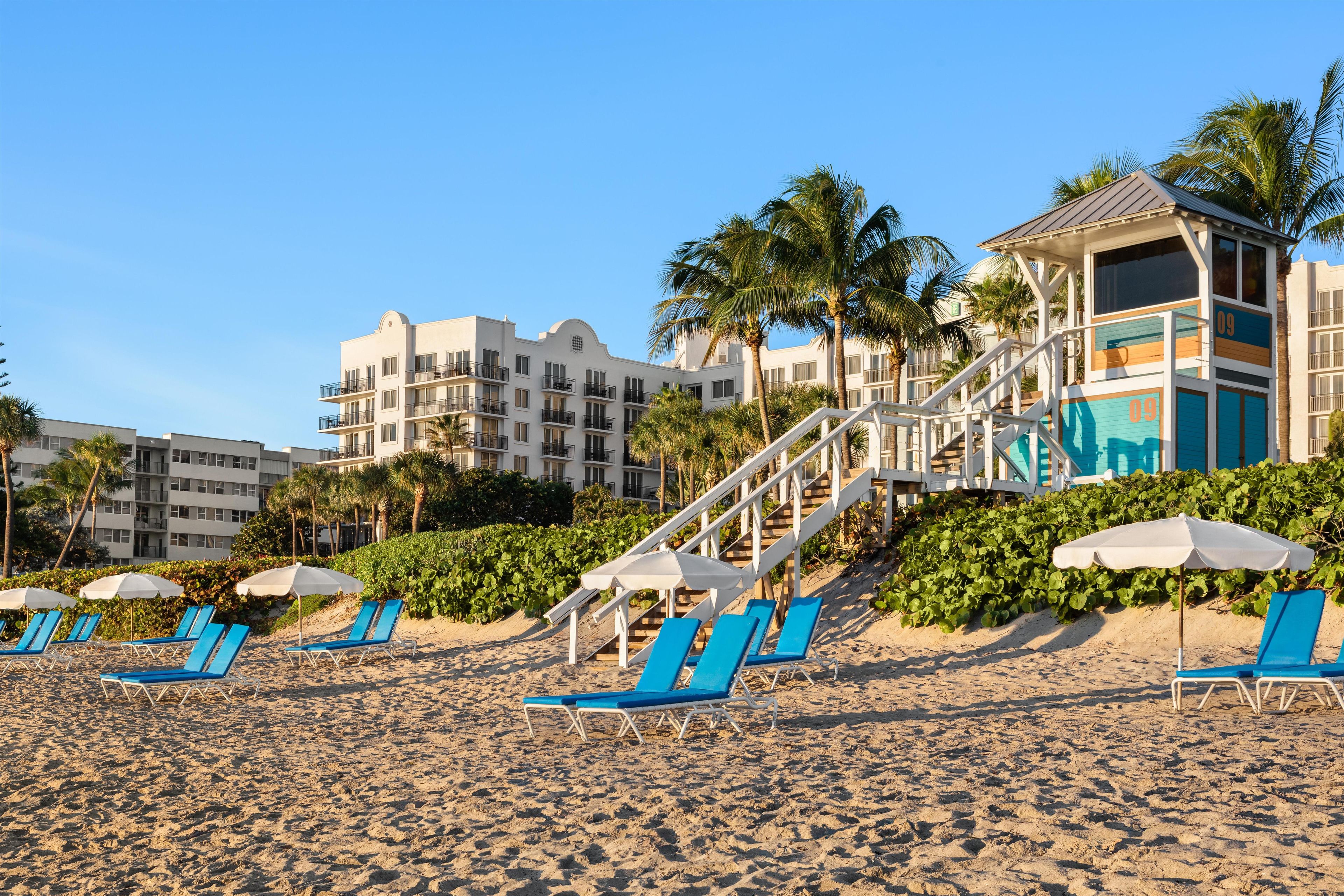 A beachfront hotel with multiple-story buildings, a staircase leading to a pool area, and several lounge chairs with umbrellas.