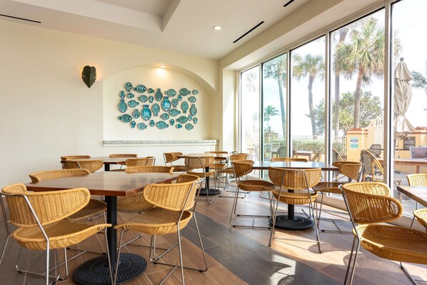 A spacious dining area with wicker chairs and tables, large windows with a view of palm trees, and a decorative wall with fish-shaped tiles.