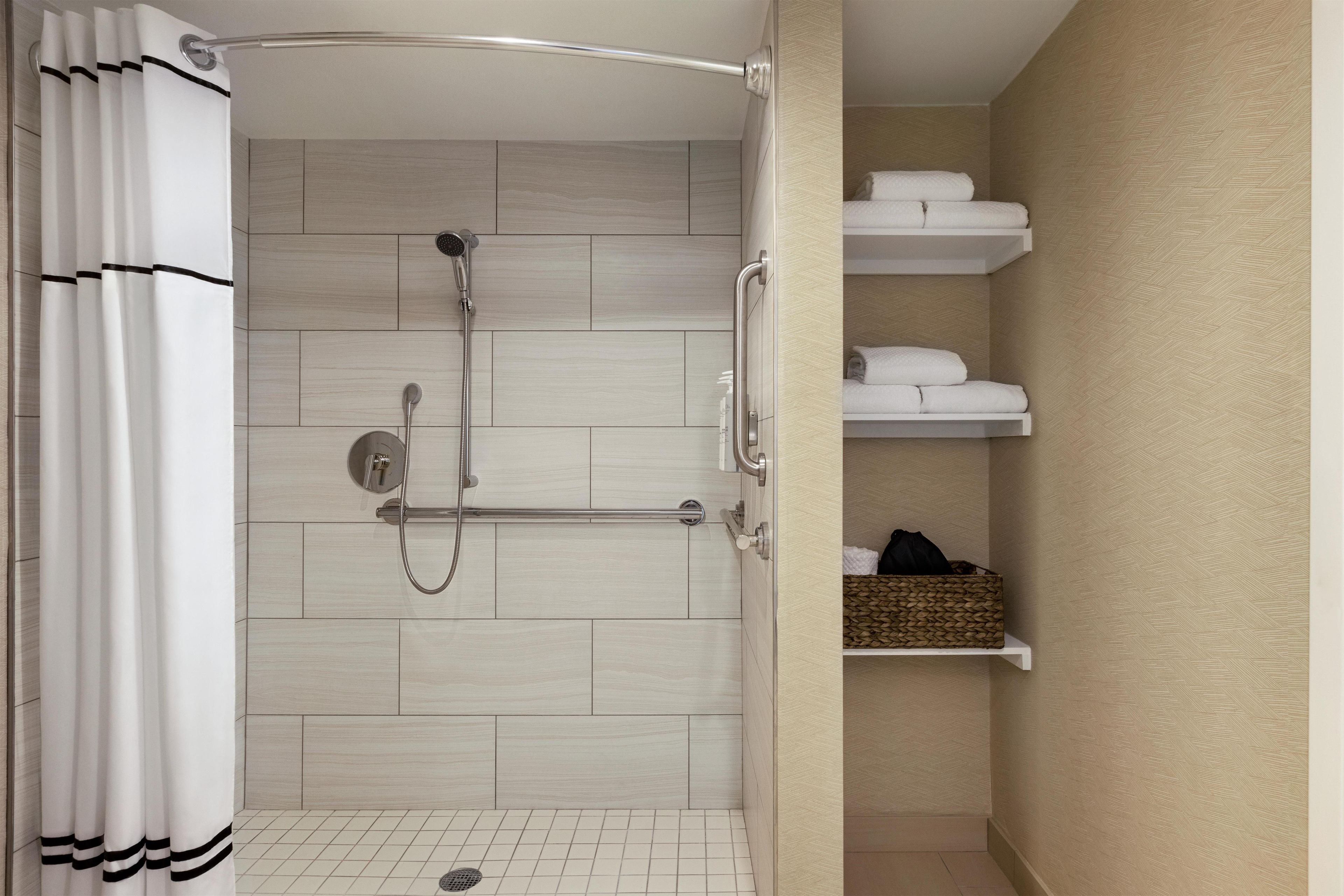 Zona de ducha con cabezal de ducha de mano, grifo empotrado en la pared y barra de apoyo. Suelos de azulejos blancos y cortina de ducha blanca con rayas negras.