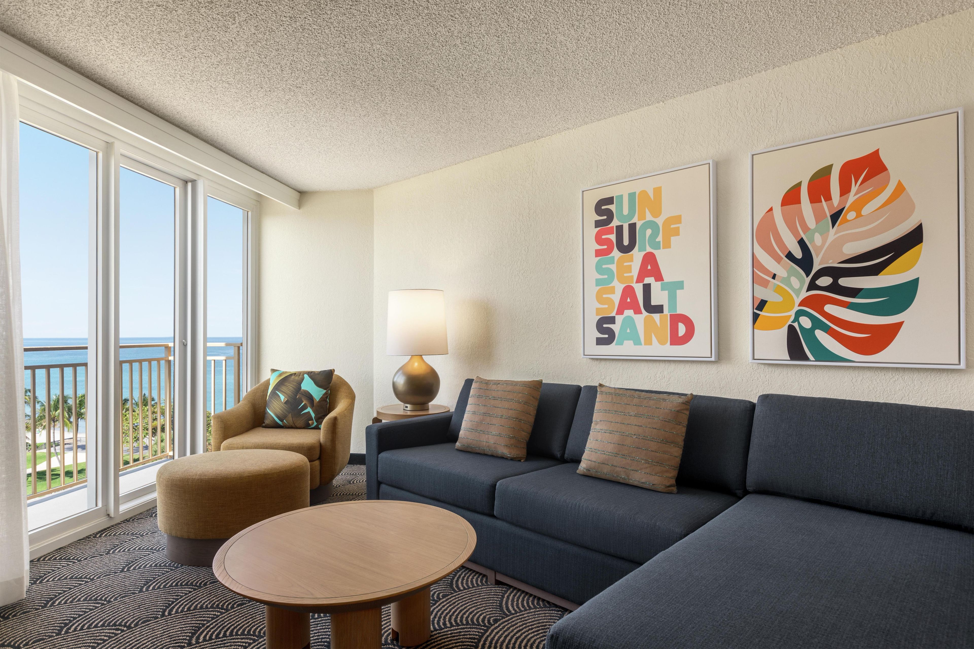 A hotel room with a sofa, armchair, round table, and artwork on the wall.
