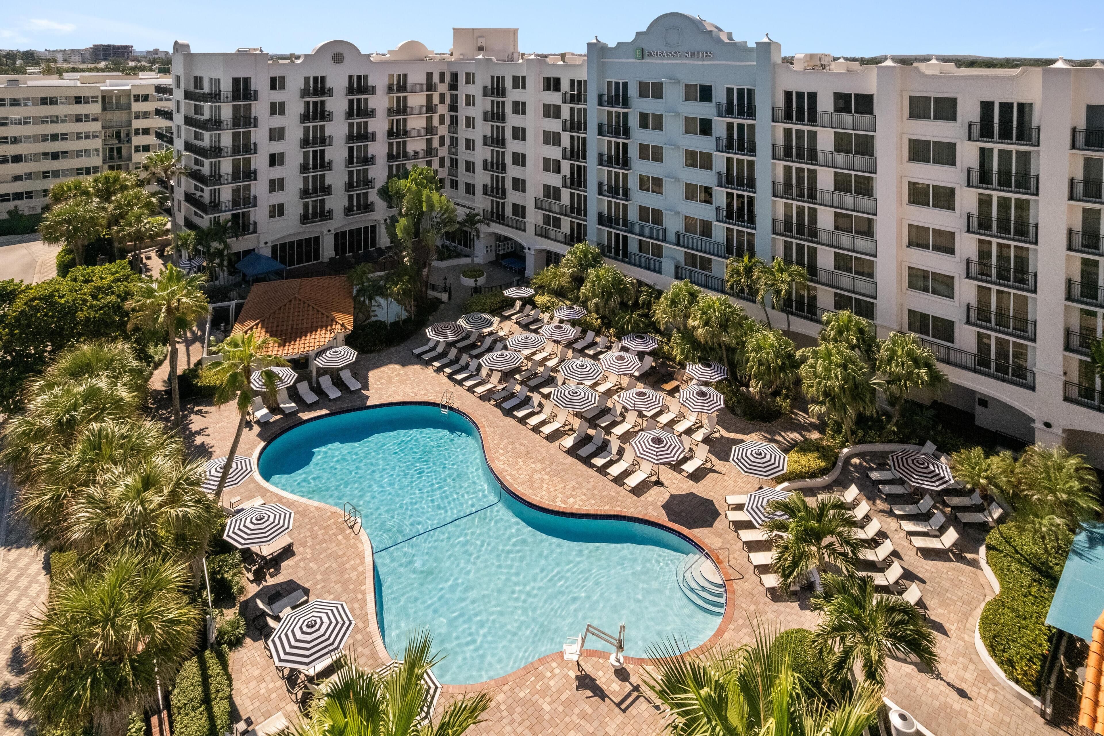 A hotel with a pool surrounded by sun loungers and umbrellas, palm trees, and a multi-story building with balconies.