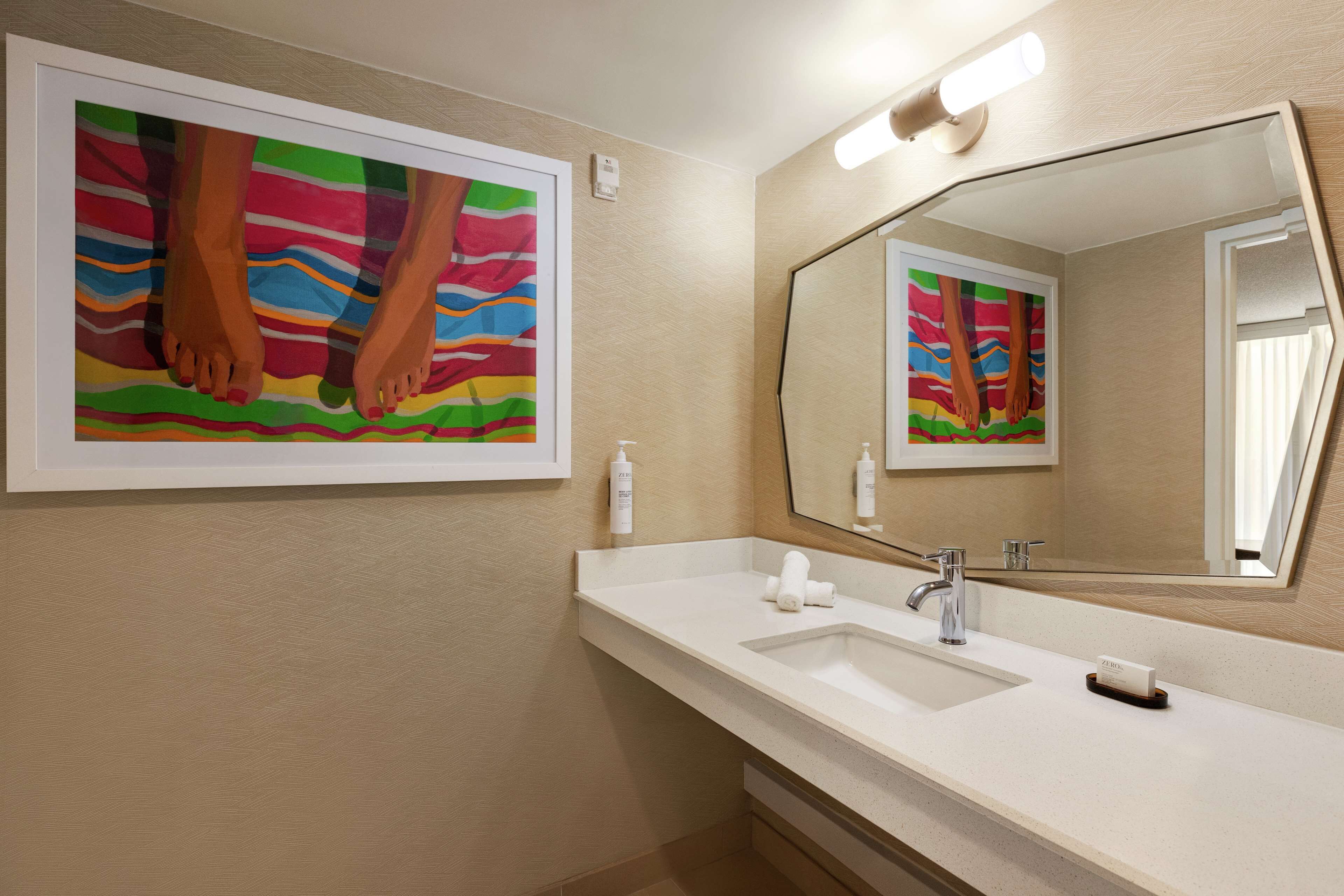 Un baño con un espejo grande, un lavamanos y una obra de arte enmarcada con la imagen de pies sobre un fondo colorido.