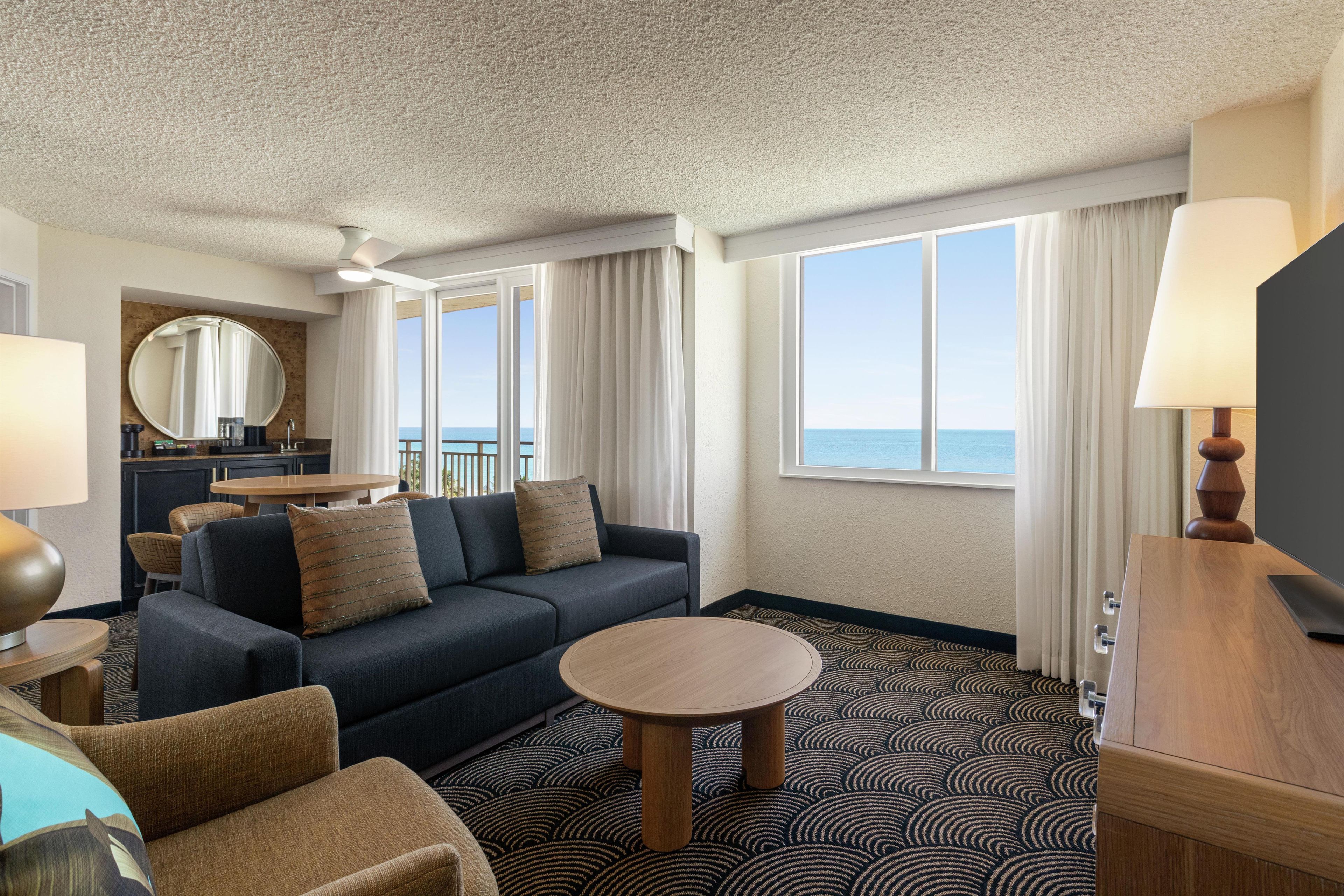 A hotel room with a sofa, armchairs, a round table, a TV, and a view of the sea.