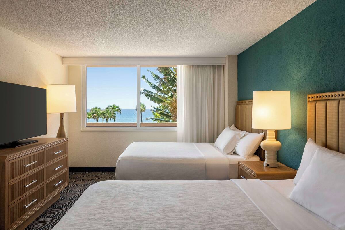 Ein Hotelzimmer mit zwei Betten, einem Fernseher und einem Fenster mit Blick auf Palmen und das Meer.
