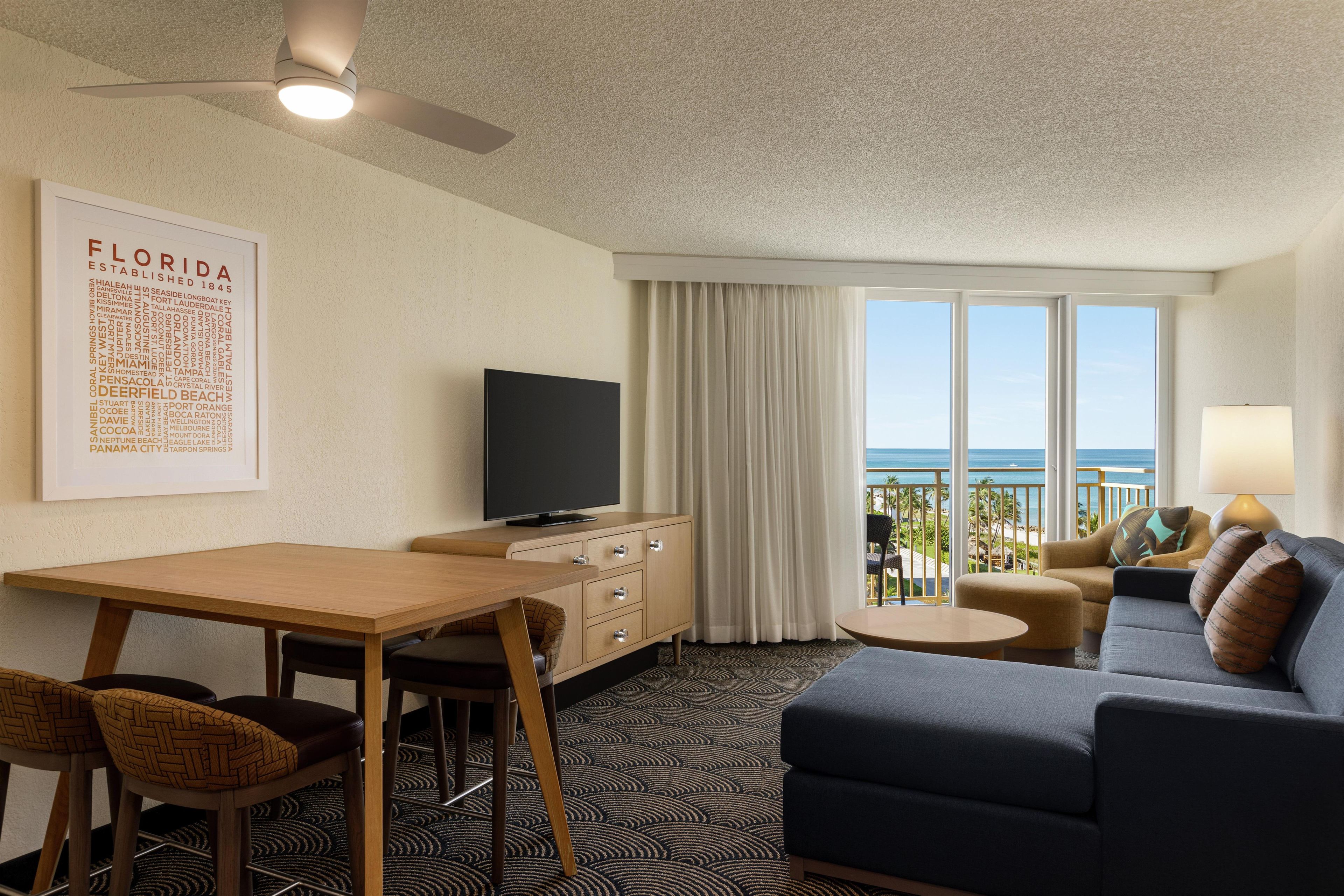 A hotel room with a flat-screen TV, a dining table, a sofa, and a balcony with a view of the ocean.