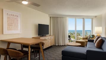 A hotel room with a flat-screen TV, a dining table, a sofa, and a balcony with a view of the ocean.