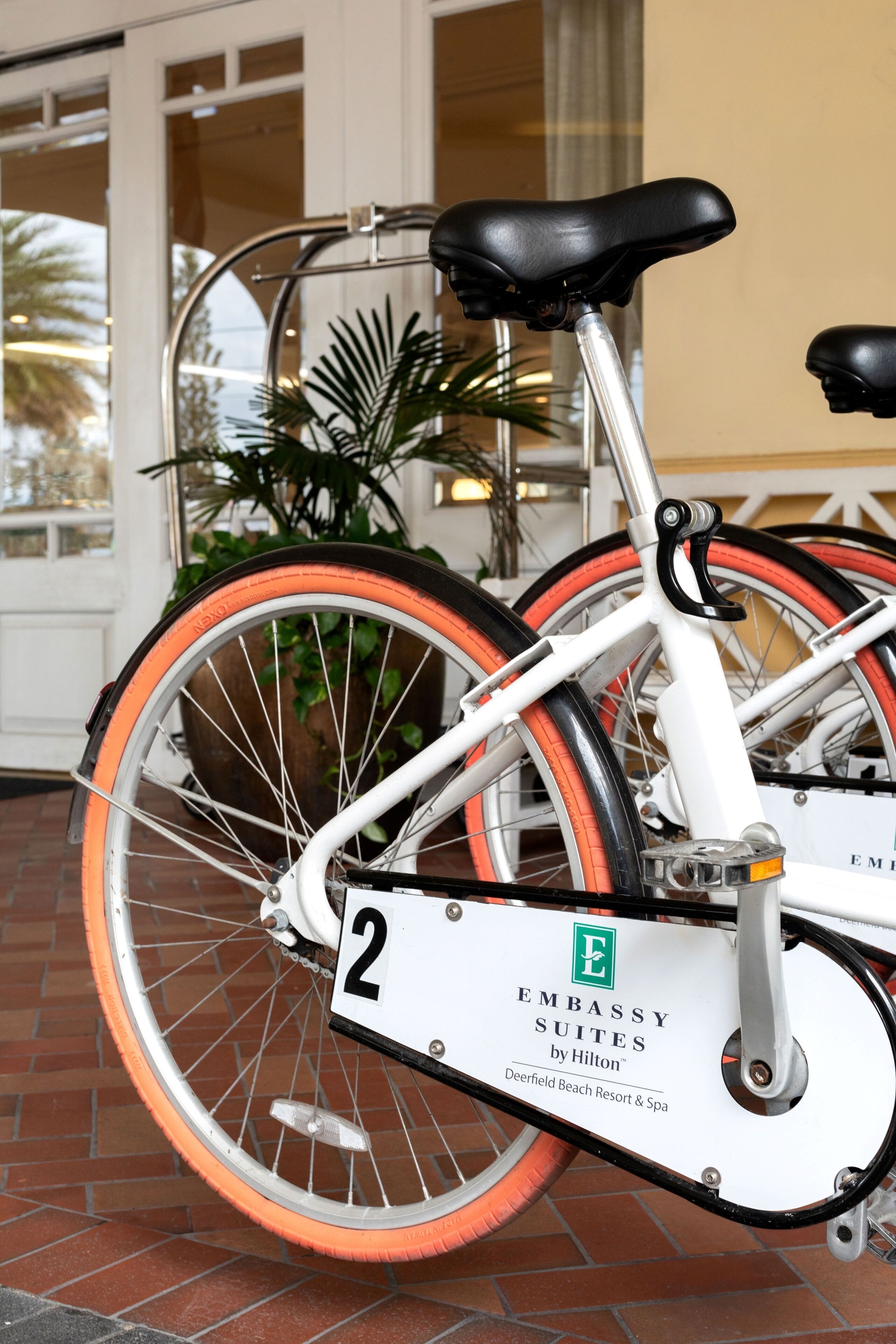 A bicycle with a white frame and orange wheels, parked in front of a building with a glass door and a sign that reads "Embassy Suites by Hilton Deerfield Beach Resort & Spa".