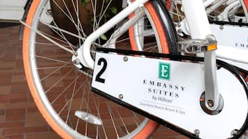 A bicycle with a white frame and orange wheels, parked in front of a building with a glass door and a sign that reads "Embassy Suites by Hilton Deerfield Beach Resort & Spa".