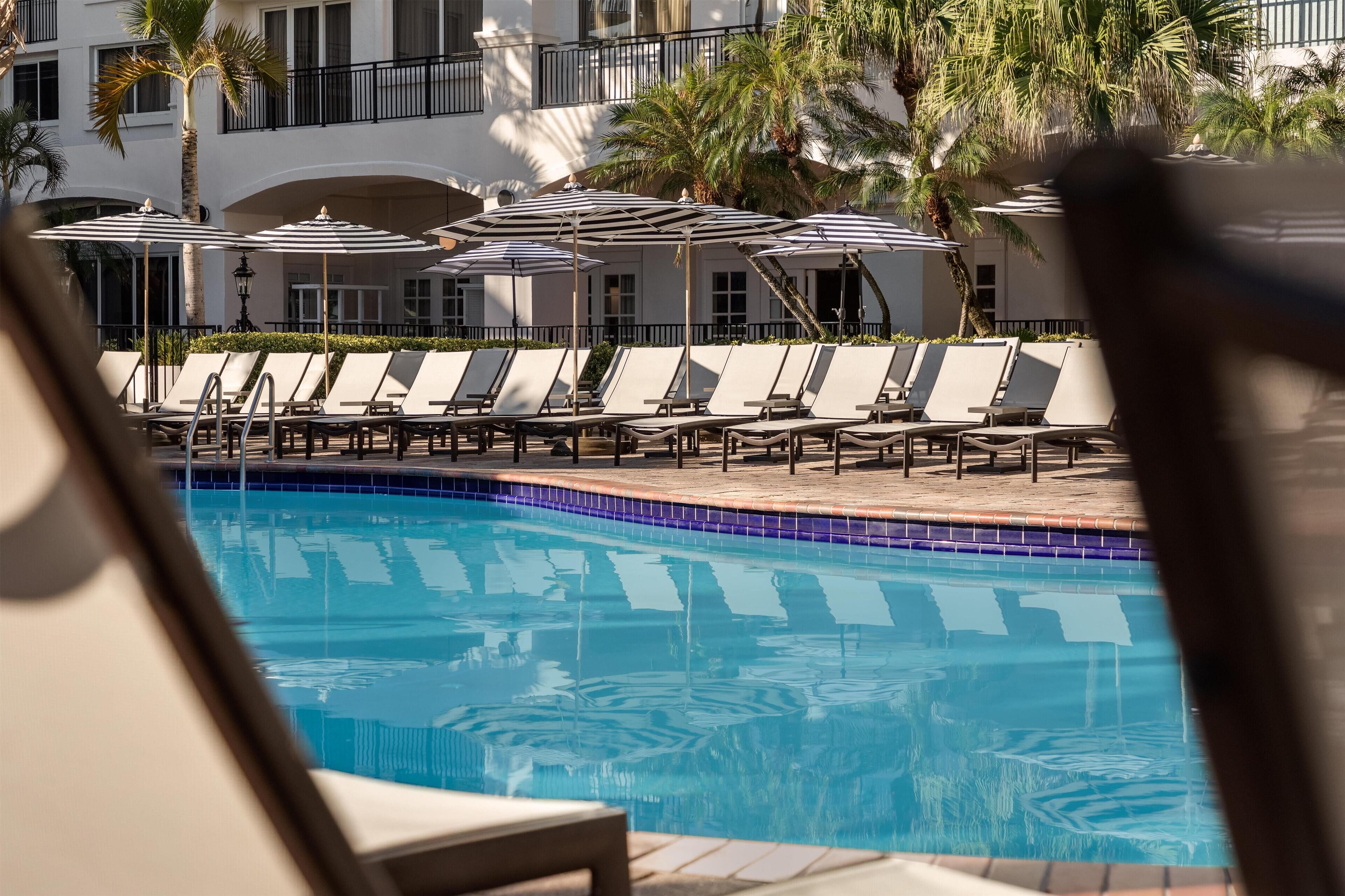 A pool area with lounge chairs and umbrellas, surrounded by palm trees and a building with balconies.