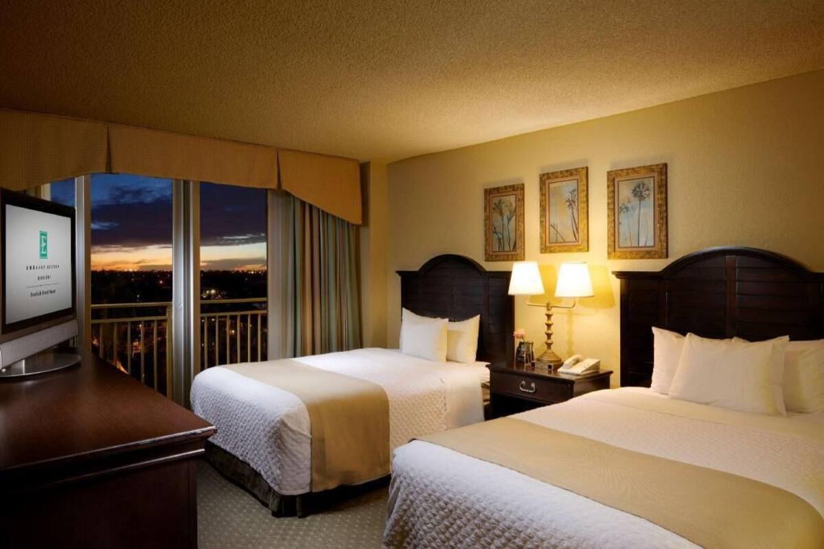 Ein Hotelzimmer mit zwei Betten, einem Schreibtisch mit Computer, einem Nachttisch mit Lampe und Blick auf die Stadt bei Abenddämmerung.