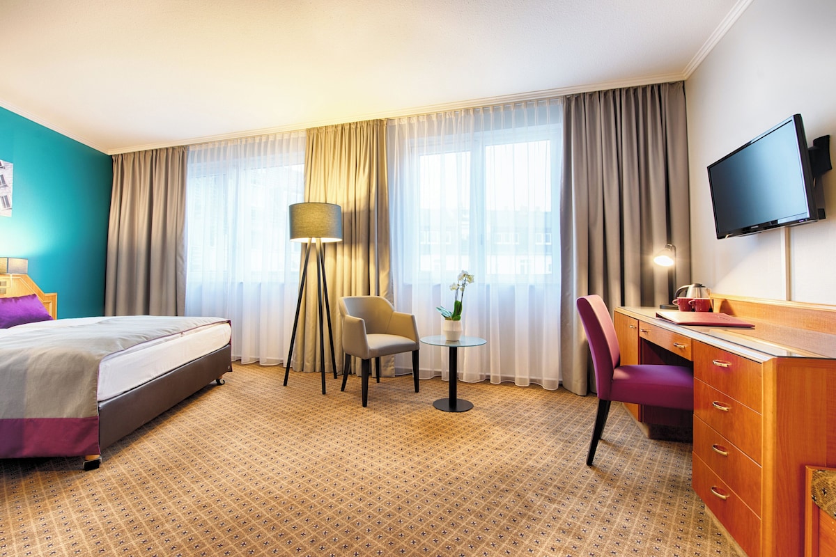 Une chambre d’hôtel avec un lit, un bureau, une chaise, une télévision et une fenêtre avec des rideaux.