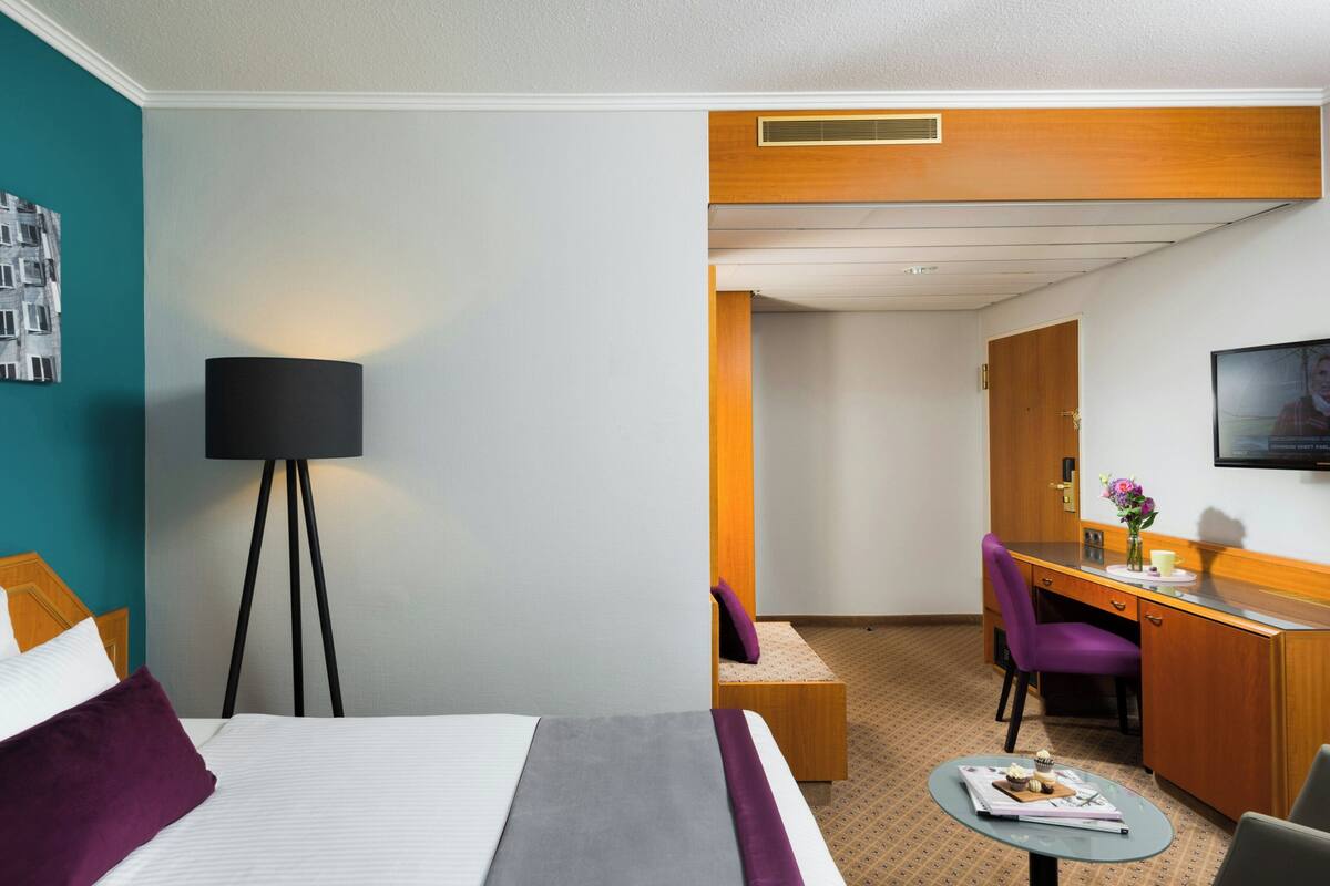 Une chambre d’hôtel avec un lit, un bureau, une chaise, une lampe et une petite table sur laquelle se trouvent une tasse et une fleur.