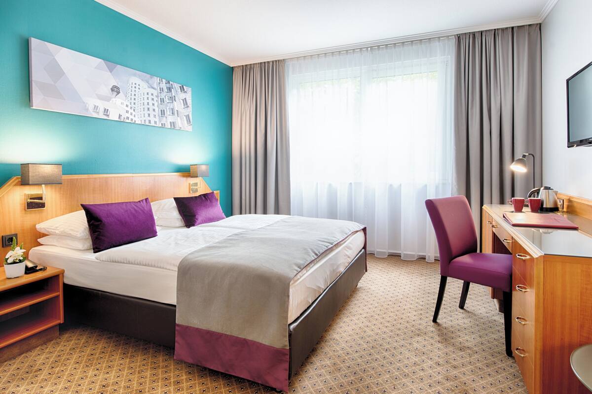 Une chambre d’hôtel avec un lit, un bureau, une chaise, une télévision et une fenêtre avec des rideaux.