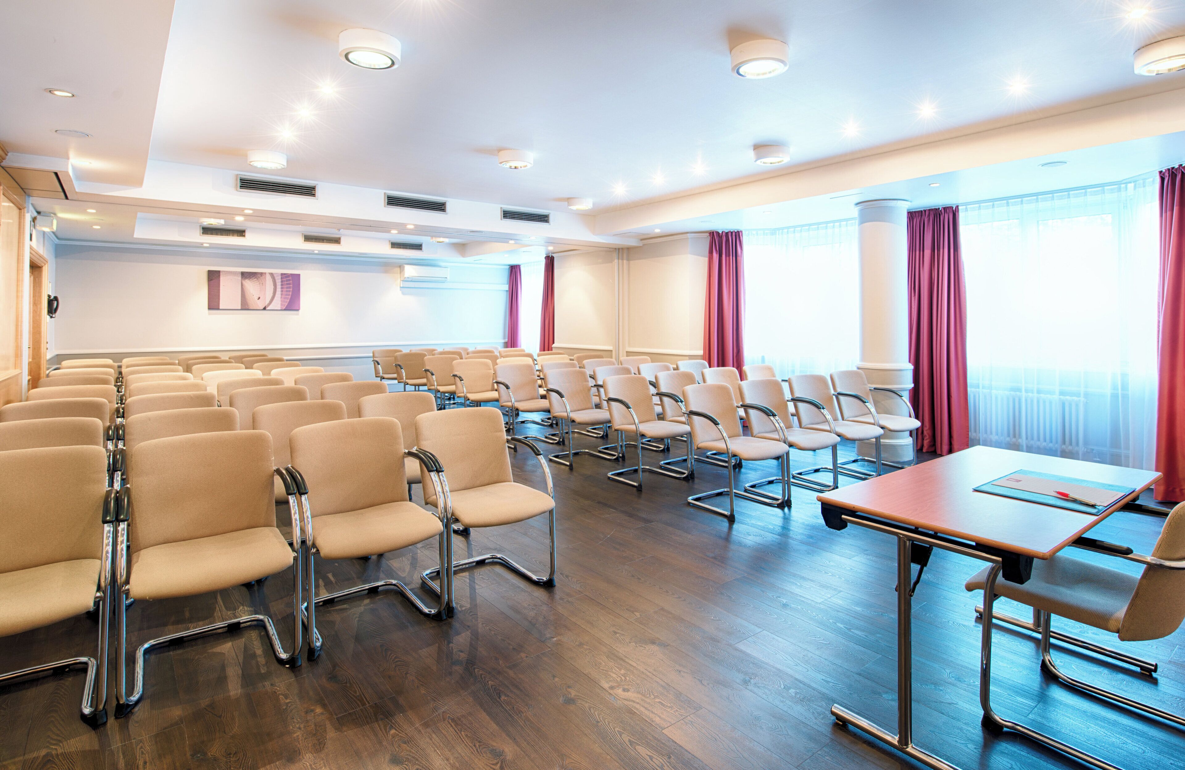 Sala de conferência com fileiras de cadeiras, uma mesa e janelas amplas com cortinas.