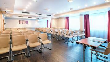 Sala de conferência com fileiras de cadeiras, uma mesa e janelas amplas com cortinas.