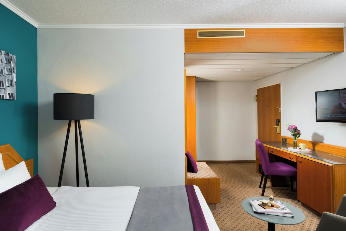 Une chambre d’hôtel avec un lit, un bureau, une chaise, une lampe et une petite table sur laquelle se trouvent une tasse et une fleur.