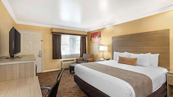 A hotel room with a large bed, a desk with a flat-screen TV, a chair, a lamp, and a window with curtains.