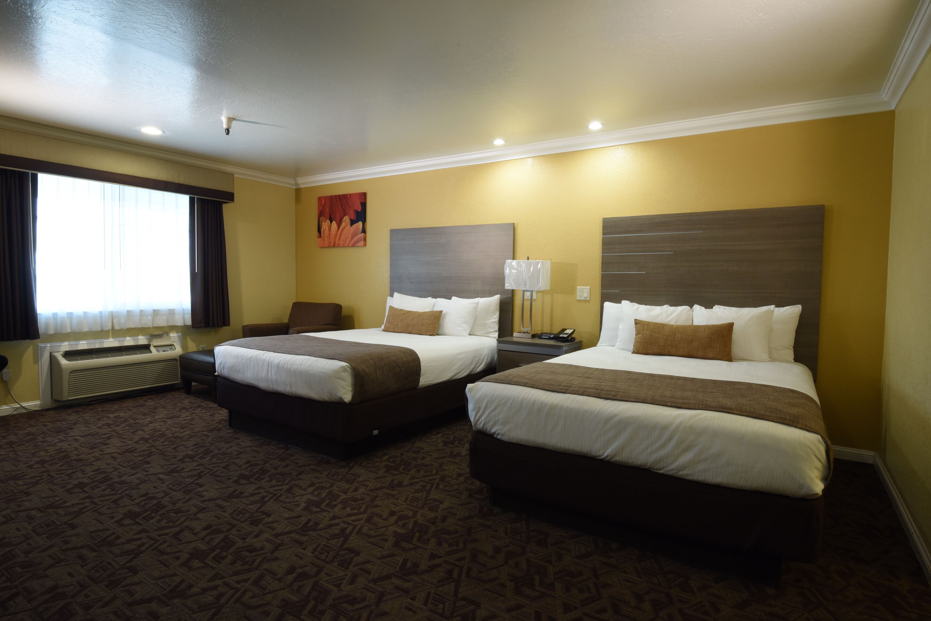 A hotel room with two beds, a desk, and a chair. There is a window with curtains, a wall-mounted heater, and a painting on the wall.